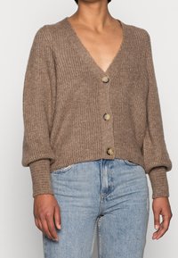 Cardigan marron en tricot avec un col en V, des manches oversize et des boutons décoratifs. Texture douce, design court, associé à un jean en denim clair.