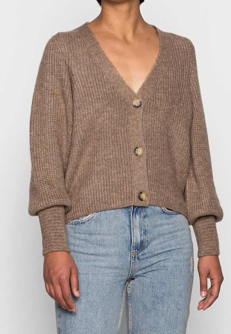Cardigan marron en tricot avec un col en V, des manches oversize et des boutons décoratifs. Texture douce, design court, associé à un jean en denim clair.