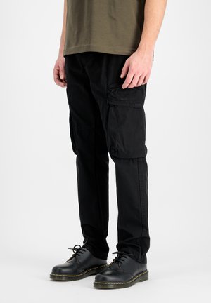 Homme portant un pantalon cargo noir avec poches à rabat et chaussures noires à lacets, debout devant un fond uni.