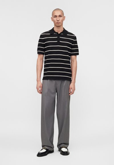 Άνδρας που φοράει κοντομάνικο πόλο με μαύρες και λευκές ρίγες, γκρι φαρδιά παντελόνια και μαύρα και λευκά μοκασίνια, στέκεται μπροστά σε απλό φόντο.