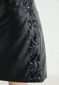 Svart skinnskjørt med sidesnøring og metalløyler, glatt tekstur og en tettsittende design. Detaljene er minimalt og stilrent.