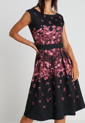 Femme portant une robe noire sans manches avec un motif de pétales floraux roses et une ceinture noire, debout avec une main sur la hanche.