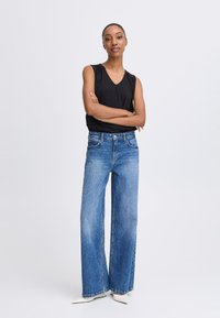 Fekete ujj nélküli felső V-nyakkal, széles lábú kék farmernadrággal párosítva. A farmernadrág standard derékkal rendelkezik és világos denim textúrájú.