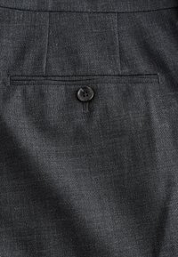 Tissu gris foncé avec un seul bouton noir retenant une poche horizontale fendue sur un vêtement taillé sur mesure.