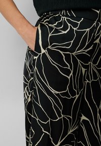Pantalons noirs avec un motif de feuilles blanches, dotés d'une taille élastique et de poches latérales. Le tissu semble lisse avec un léger brillant.