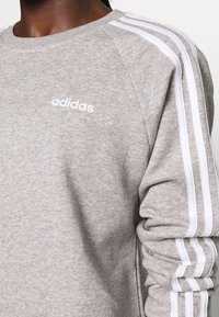 Persona que lleva un sudadera Adidas de un gris claro con detalles de tres franjas blancas en las mangas y el logo de Adidas en el pecho.