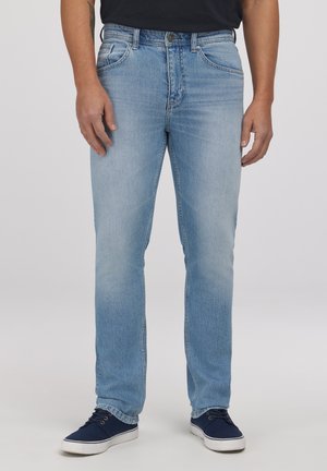 Jasnoniebieskie jeansy o prostych nogawkach noszone przez stojącą osobę, zestawione z granatowymi sportowymi trampkami na jednolitym tle.