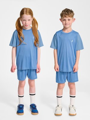 Twee kinderen staan naast elkaar in bijpassende lichtblauwe sportkleding, witte sokken met zwarte strepen en sportschoenen op een effen achtergrond.