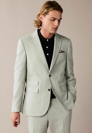 Chaqueta de traje gris claro con diseño de un solo botón, solapas con muesca y bolsillos laterales. Llevada sobre una camiseta tipo polo negra con botones.