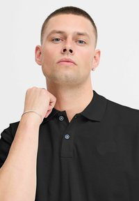 Sort polo shirt med struktureret stof, der har en klassisk krave og en to-knaps knaplukning, prydet med blå knapper. Sølvarmbånd synligt på håndleddet.