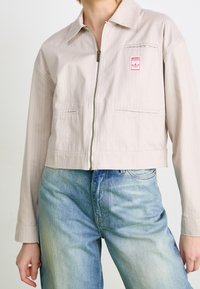 Cropped, light beige jacket with a striped pattern, front zipper, and two side pockets. Features a small pink logo patch on the breast.