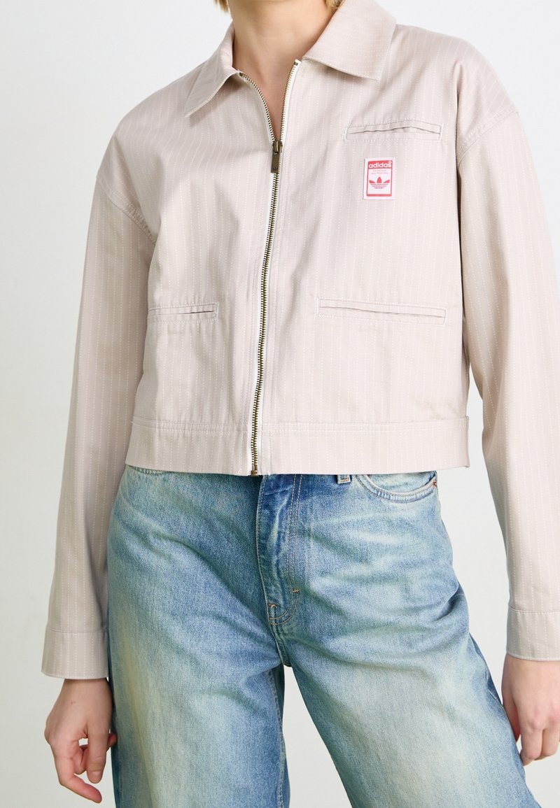 Cropped, light beige jacket with a striped pattern, front zipper, and two side pockets. Features a small pink logo patch on the breast.