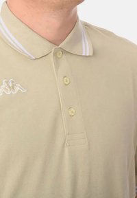 Polo beige con tre bottoni e colletto bicolore con strisce bianche; presenta un logo ricamato sul lato sinistro del petto.