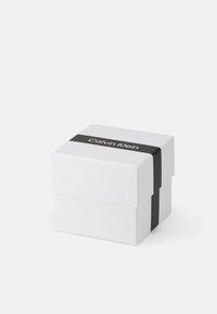 Caja de regalo blanca en forma de cuadrado con una banda de papel negra que presenta el texto "Calvin Klein". Textura suave, diseño minimalista.