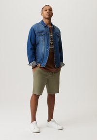 Jeansowa kurtka z jaśniejszym niebieskim korpusem i ciemnoniebieskimi rękawami, brązowy T-shirt z grafiką, oliwkowe szorty i białe trampki.