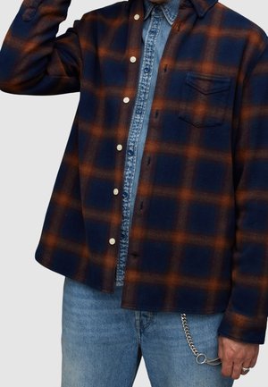 Chemise à carreaux bleu marine et orange avec un devant boutonné, une poche poitrine et des manches longues, superposée à un haut en denim bleu clair.