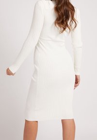 Femme portant une robe blanche à manches longues et côtelée, se tenant de dos face à la caméra devant un fond uni.