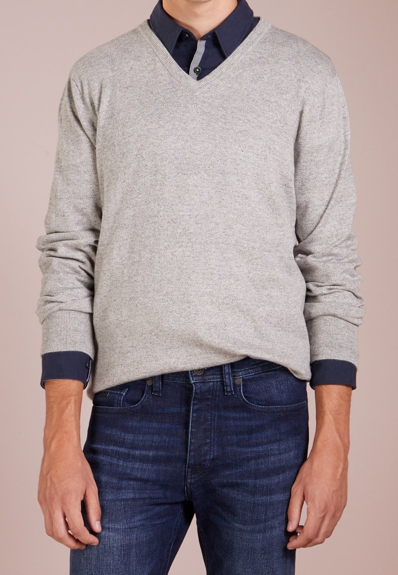 Homme portant un pull gris clair à col en V par-dessus une chemise bleu marine à boutons avec un jean bleu foncé, mains détendues le long du corps.