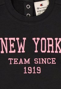 Black sweatshirt featuring pink embroidered text: "NEW YORK TEAM SINCE 1919" with snap button closure at the neck and a soft texture.