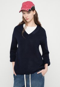 Μπλε ναυτικό πουλόβερ με V λαιμόκοψη φτιαγμένο από μαλακό υλικό, με ρεβέρ στα μανίκια και ραβδωτή ρόμβωση στο τελείωμα. Συνδυασμένο με ροζ καπέλο και casual τζιν παντελόνια.