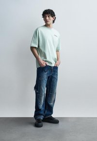 Jeune homme aux cheveux bouclés portant un T-shirt oversize vert clair, un jean bleu foncé large avec des patchs, et des baskets noires, debout les mains dans les poches.