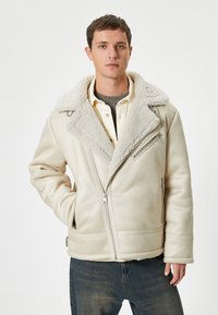 Krämfärgad fårskinnsjacka med bred krage, sneddragkedjor och mjuk textur, ackompanjerad av en ljus skjorta och mörka jeans.