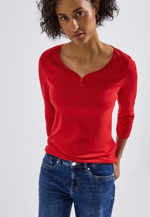 Rode langemouwen shirt met een ronde halslijn en drie knopen bij de sluiting, gecombineerd met een blauwe jeans met een klassieke pasvorm.