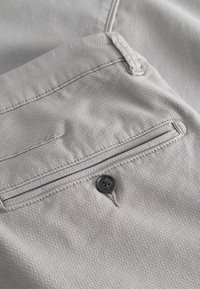 Tela de color gris claro con un tejido texturizado que presenta un bolsillo trasero con un botón negro y bordes cosidos.