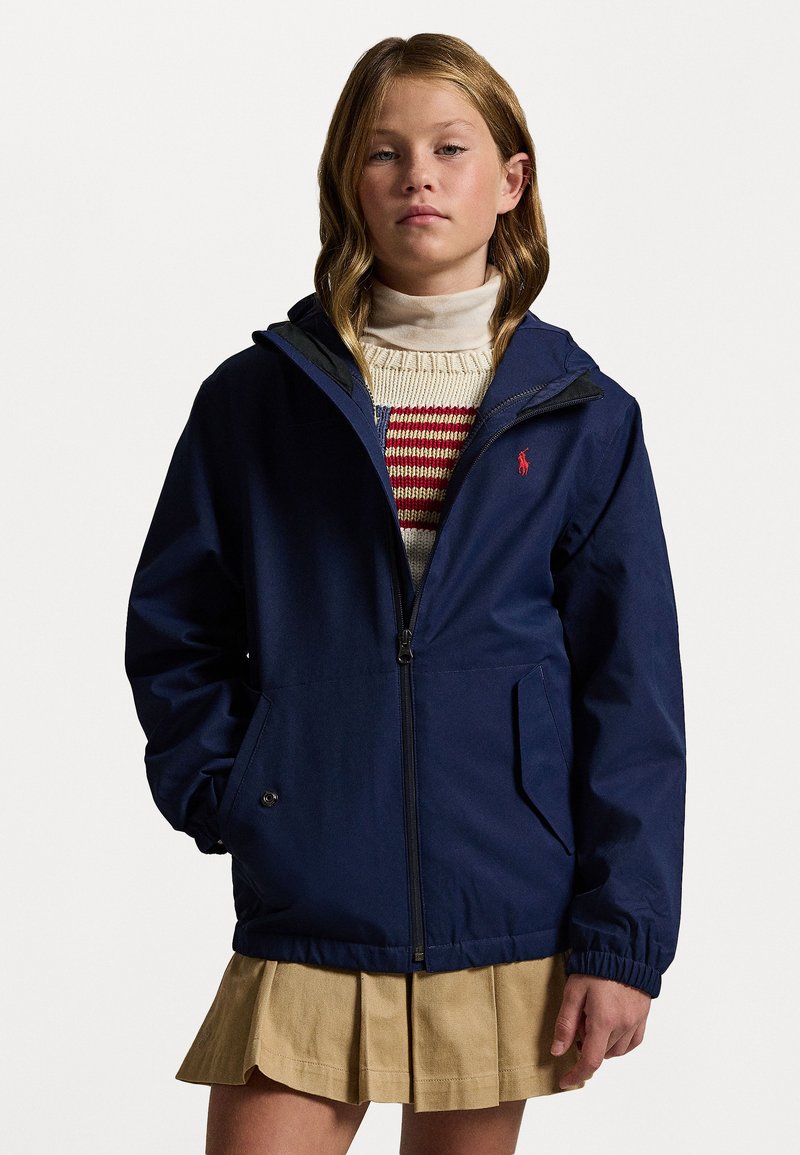 Marineblaue Zip-Jacke mit Kapuze und elastischen Bündchen, mit einem kleinen roten Logo. Getragen über einem cremefarbenen Pullover mit roten Streifen und einem beigefarbenen Faltenrock.