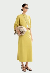 Sweatshirt jaune coupé associé à une jupe longue assortie. Le mannequin est accessoirisé avec des lunettes de soleil noires et un collier en pendentif argenté.
