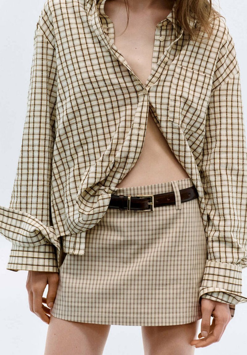 Femme portant une chemise à carreaux beige et une jupe assortie à carreaux avec une ceinture marron foncé, montrant le ventre, debout devant un fond uni.