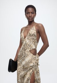 Γυναίκα που φοράει αμάνικο φόρεμα με μοτίβο φίδι και ανοίγματα, κρατώντας μια μαύρη τσάντα χειρός μπροστά σε απλό φωτεινό φόντο.