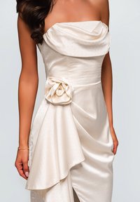 Abito in satin avorio senza spalline con corpetto drappeggiato, accentuato da un dettaglio floreale in vita e una delicata texture a pois. Finitura liscia e orlo asimmetrico.