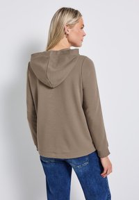 Felpa color beige con una vestibilità comoda, realizzata in materiale morbido, con un yoke posteriore e un cappuccio. Abbinata a jeans di denim blu.