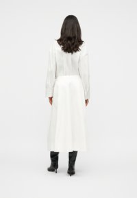 Femme aux longs cheveux noirs portant une chemise blanche à rayures manches longues, une jupe midi blanche et des bottes à talons noires, debout de dos.