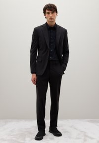 Uomo in piedi con le mani in tasca, che indossa un completo nero attillato, una camicia nera e scarpe slip-on nere, su uno sfondo semplice.