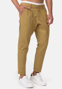 Pantalones caqui ajustados con pliegues, bolsillos delanteros y cierre de botón. Combinados con zapatillas blancas con acentos negros.