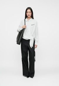 Blusa blanca con detalle de lazo y logo, combinada con pantalones negros de pierna ancha que presentan cremalleras y tirantes. Modelo sosteniendo un bolso negro.