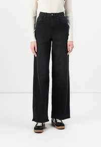High-waisted black denim jeans with wide legs and front pockets. Features a button closure and shown paired with black sneakers.