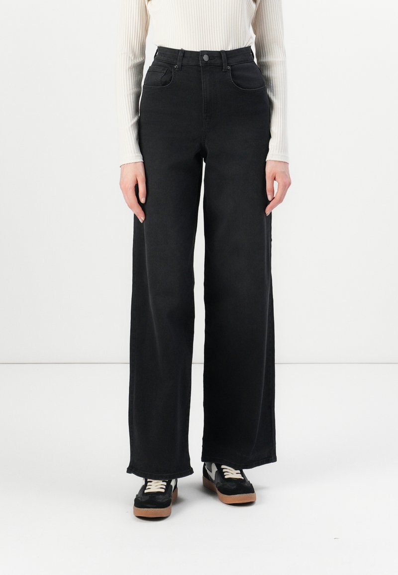High-waisted black denim jeans with wide legs and front pockets. Features a button closure and shown paired with black sneakers.