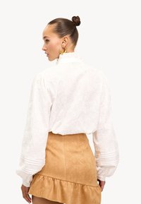 Witte blouse met ingewikkelde textuur en ruches kraag, gecombineerd met een tan kleurige suède rok met een gefronste zoom. Gouden oorringen zichtbaar.