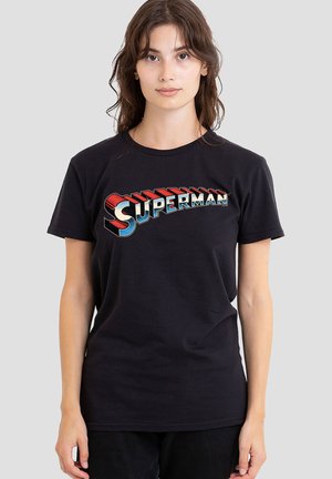 Young woman with brown hair wears a black t-shirt with a colorful, retro-style "Superman" logo on the chest against a plain background.
