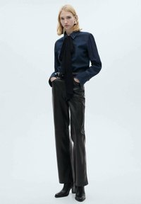 Camicia di denim con dettaglio cravatta nera, abbinata a pantaloni in pelle nera a vita alta e stivaletti, esposta su uno sfondo neutro.