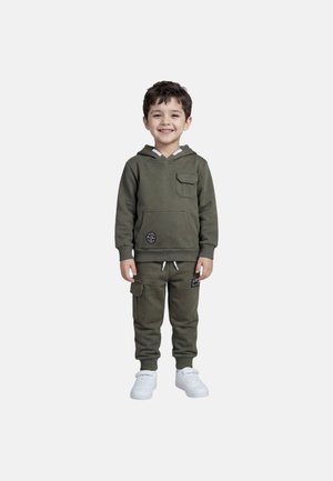 Jeune garçon souriant debout, portant un sweat à capuche vert olive et un pantalon de jogging assorti avec des baskets blanches, sur un fond blanc uni.