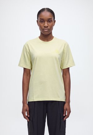 Une femme se tient face à l’objectif, vêtue d’un T-shirt jaune pâle à manches courtes et d’un pantalon noir, sur un fond clair et uni.