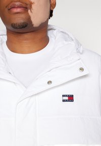 Hvid hættejakke lavet af letvægtsstof, med en lynlåslukning foran, trykknapper og et flerfarvet logo-patch på venstre bryst.