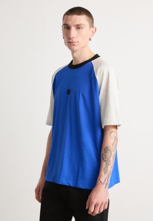 Blue short-sleeve t-shirt with gray raglan sleeves, black collar, and a center front black dot detail. Soft texture and relaxed fit.
