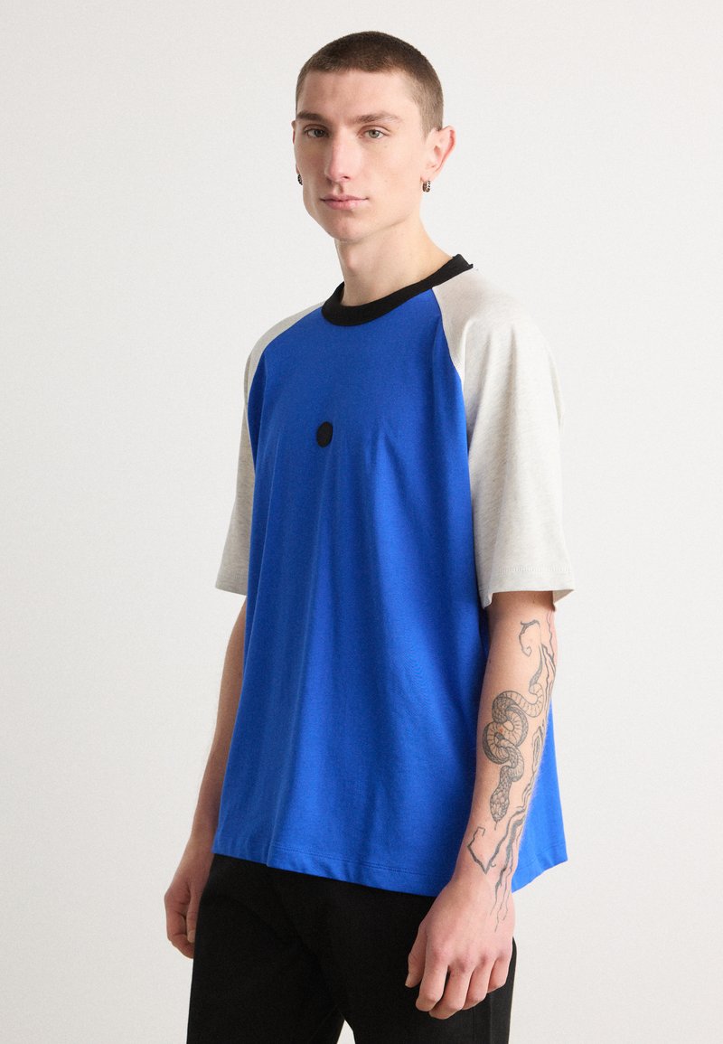 Blue short-sleeve t-shirt with gray raglan sleeves, black collar, and a center front black dot detail. Soft texture and relaxed fit.