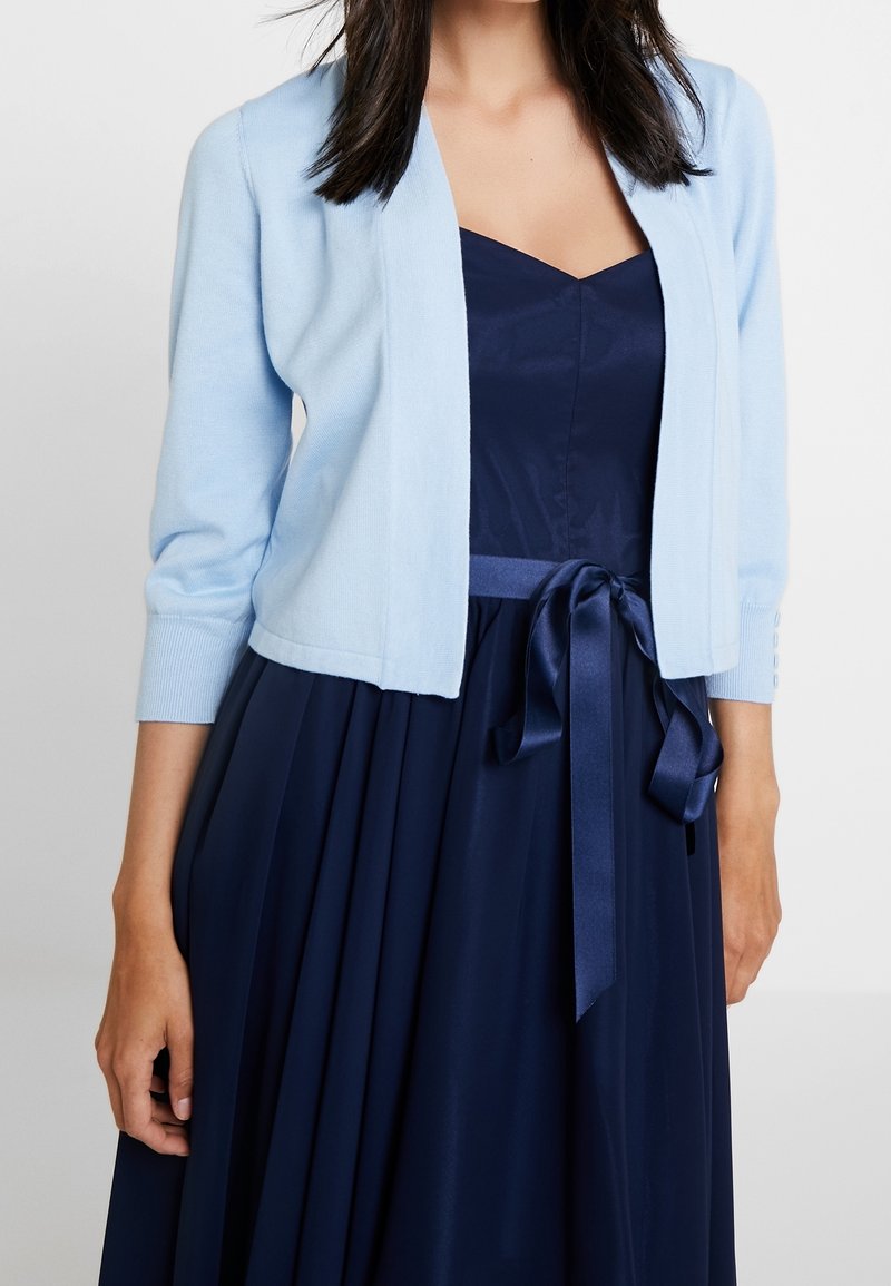 Femme portant une robe bleu marine avec une ceinture en satin à la taille et un cardigan court bleu clair à manches trois-quarts.