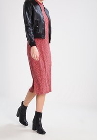 Černá kožená bomber bunda přes červené vzorované midi šaty s bočními rozparky, doplněné černými kotníkovými botami na podpatku. Hladké textury.